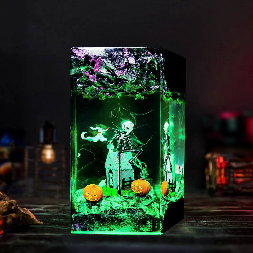 A glowing rectangular resin candle or decorative box with a Halloween scene inside, featuring a skeleton standing in front of a haunted house, green mist effects, orange pumpkins, and a dark tree with purple leaves. The item is photographed on a dark tabletop with blurred ambient lights in the background.