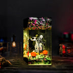 A rectangular clear acrylic aquarium-style decorative piece with a Halloween-themed scene inside, including a skeleton figure, small house, orange pumpkins, and colorful fish decorations on the water surface, photographed against a dark blurred background.