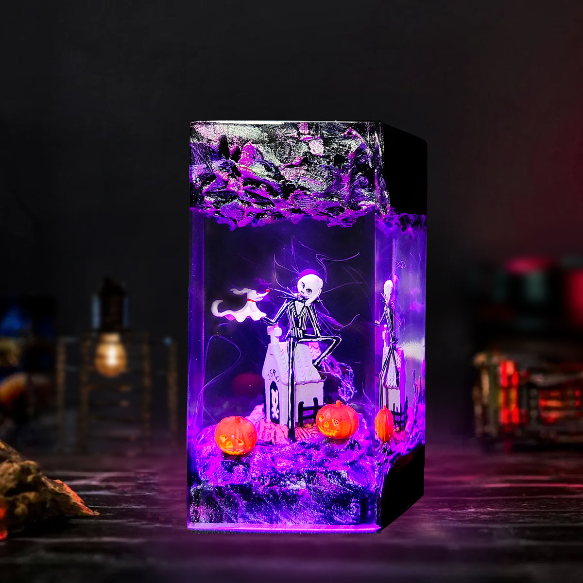 A rectangular clear resin block night light featuring a skeleton wizard sitting on a small chair, purple misty effects, two orange pumpkins, and a black side base, photographed on a dark tabletop with blurred ambient background lights.