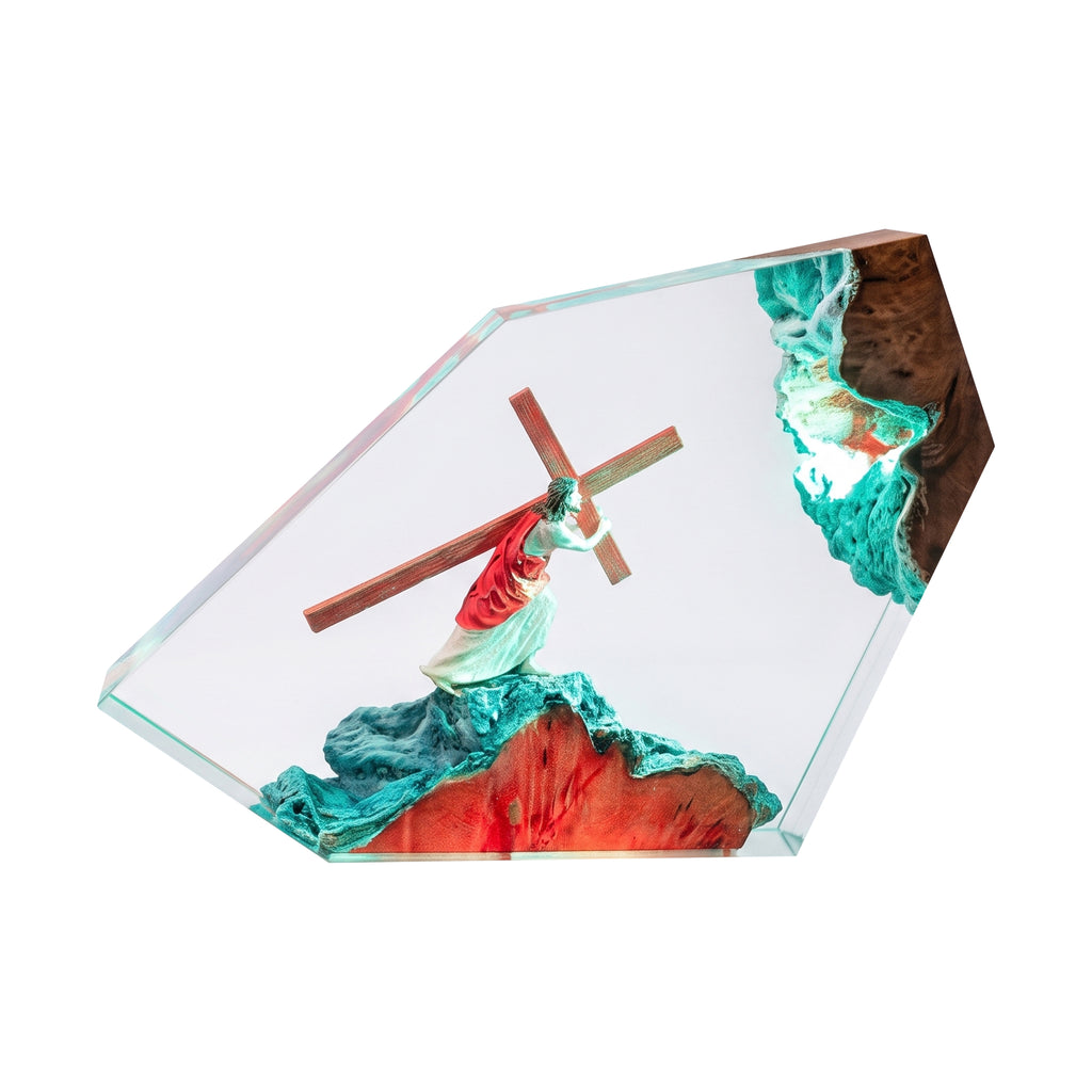 A clear resin art plaque featuring a figurine of Jesus carrying a wooden cross, set against a textured background with teal, red, and brown details, all enclosed in a geometric clear acrylic frame on a white background.