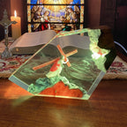 A clear resin geometric night light featuring a stained-glass-style depiction of Jesus carrying the cross, shown lit on a wooden table with a candle, open book, and stained-glass window in the background.