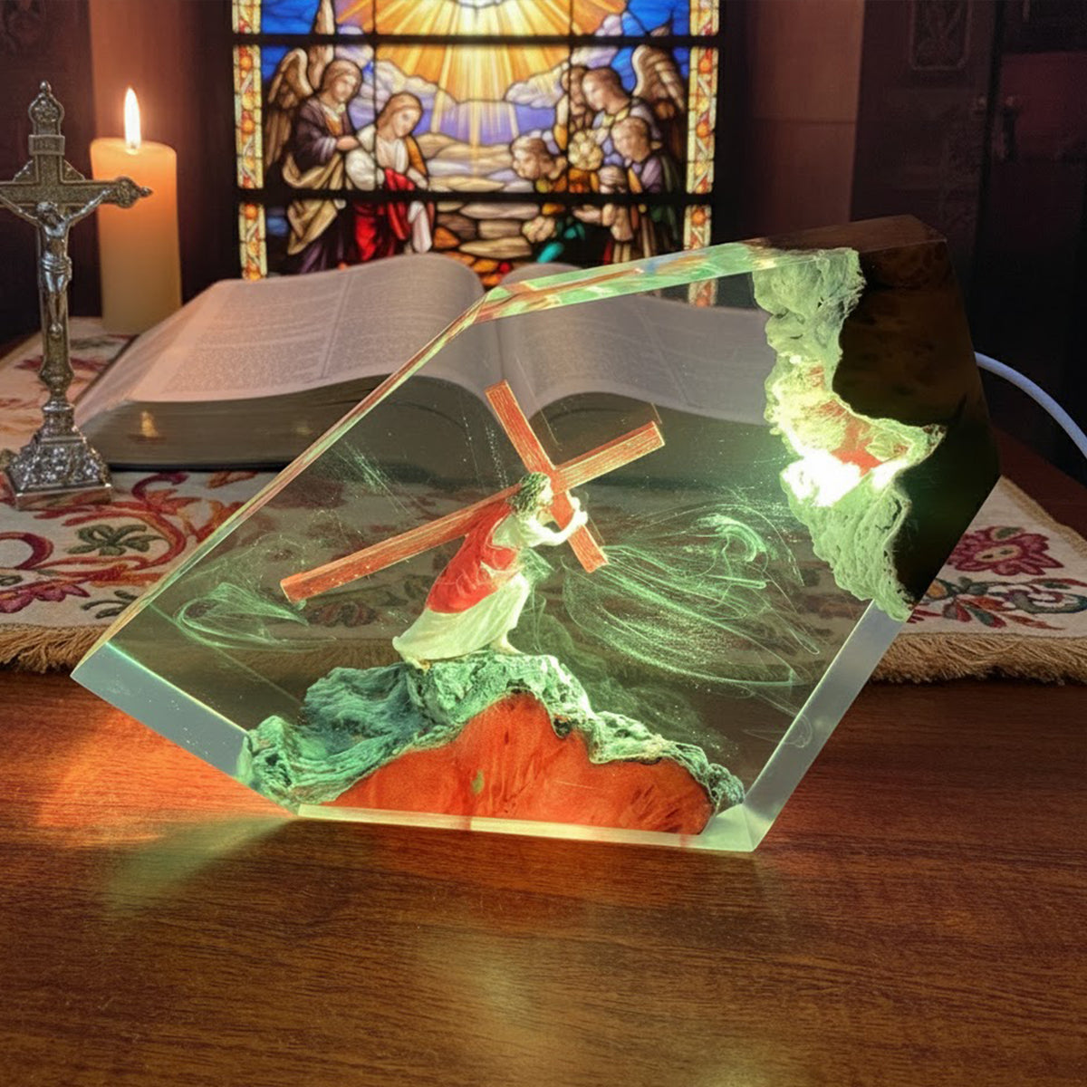 A clear resin geometric night light featuring a stained-glass-style depiction of Jesus carrying the cross, shown lit on a wooden table with a candle, open book, and stained-glass window in the background.