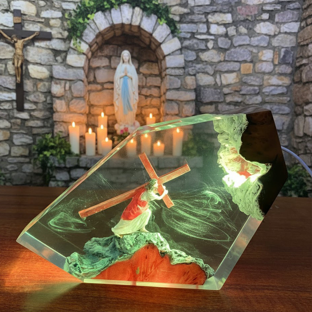 A hexagonal clear resin lamp featuring a miniature Jesus carrying the cross on a green rocky hill with glowing internal lights, displayed on a wooden surface in front of a stone shrine with candles.