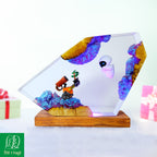 A clear acrylic resin art piece shaped like a faceted crystal, resting on a rectangular wooden base. The resin contains colorful embedded rock formations in purple, blue, and gold, along with tiny figurines including a small astronaut and a tiny rocket ship.