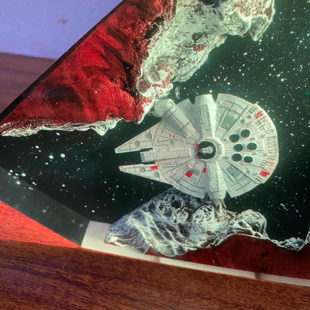 A close-up of a decorative glass plate featuring a printed or embedded scene of the Millennium Falcon floating in space above a red planet and a white moon, with a wooden surface visible around it.