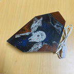 A Star Wars themed LED light shaped like a spaceship cockpit, featuring a Millennium Falcon on a space background with brown wood-grain details, shown on a light wood tabletop next to its power cable.