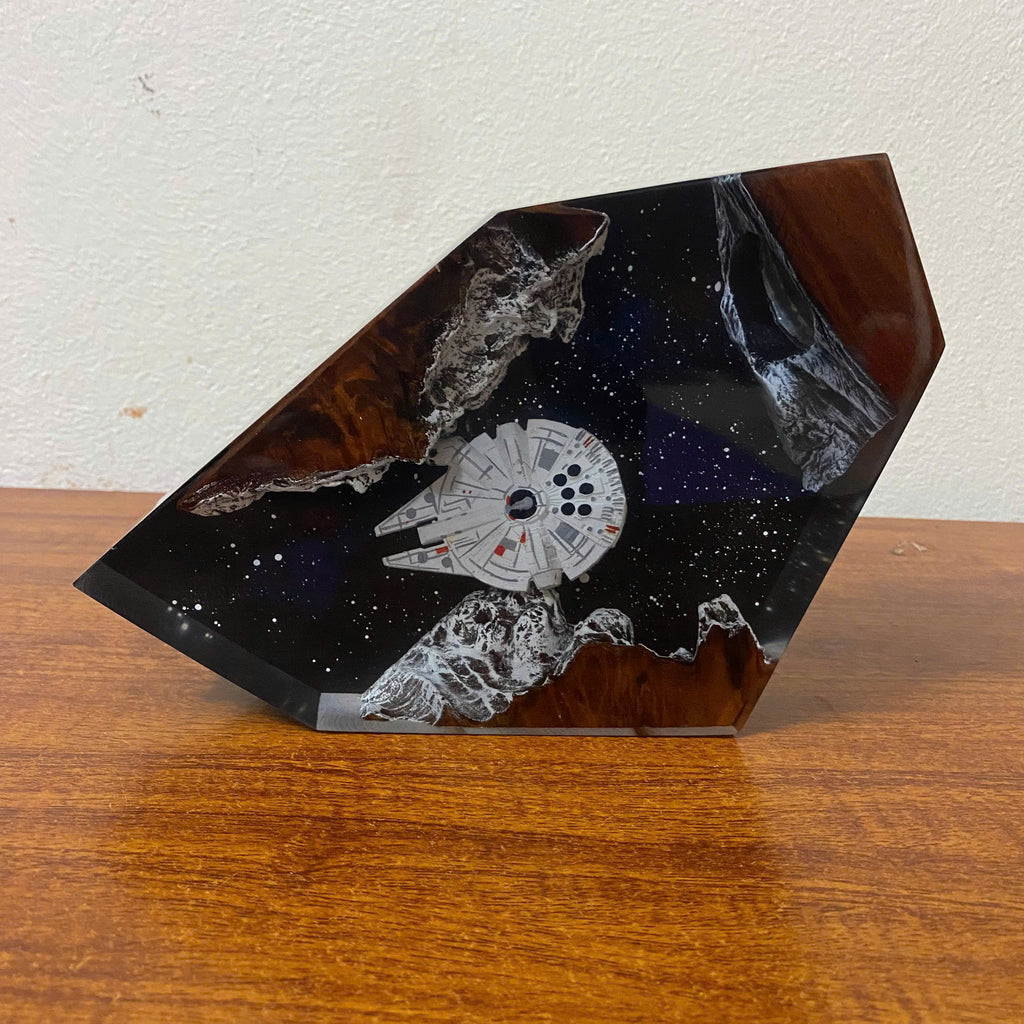 A custom resin tabletop sculpture shaped like a geometric shard, featuring a white Millennium Falcon spaceship embedded in a black starry resin, with brown wood-like swirls and white textured rock details, displayed on a wooden table against a white wall.