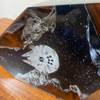 A close-up of a decorative resin or glass object featuring a white Millennium Falcon spaceship embedded in a dark, starry space design with gray rocky formations, resting on a wooden surface.
