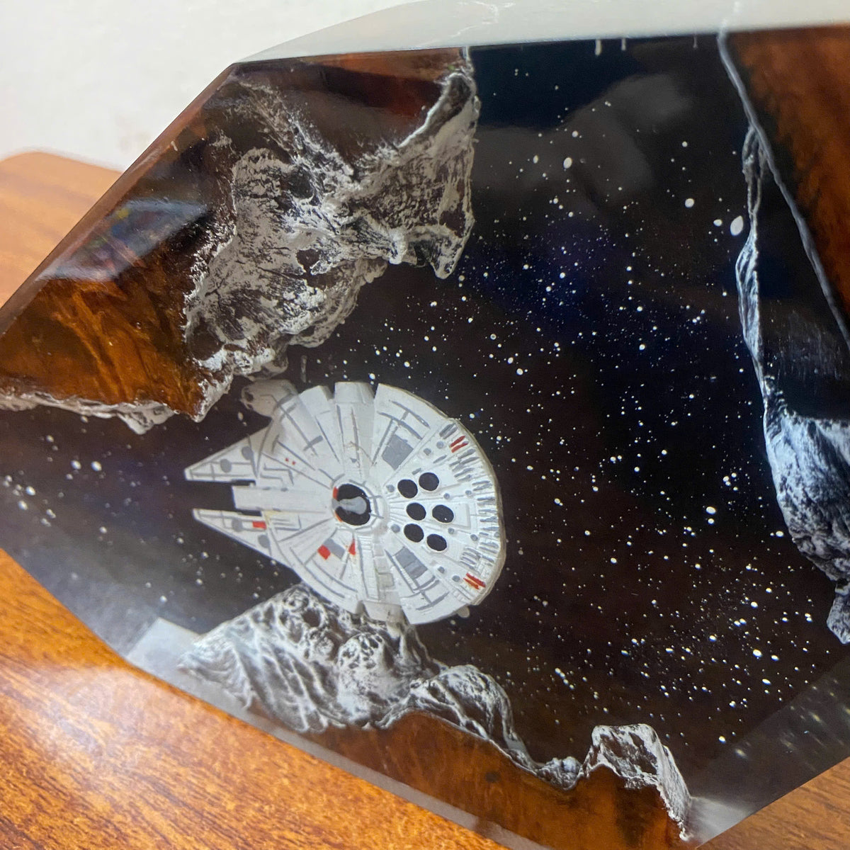 A close-up of a decorative resin or glass object featuring a white Millennium Falcon spaceship embedded in a dark, starry space design with gray rocky formations, resting on a wooden surface.