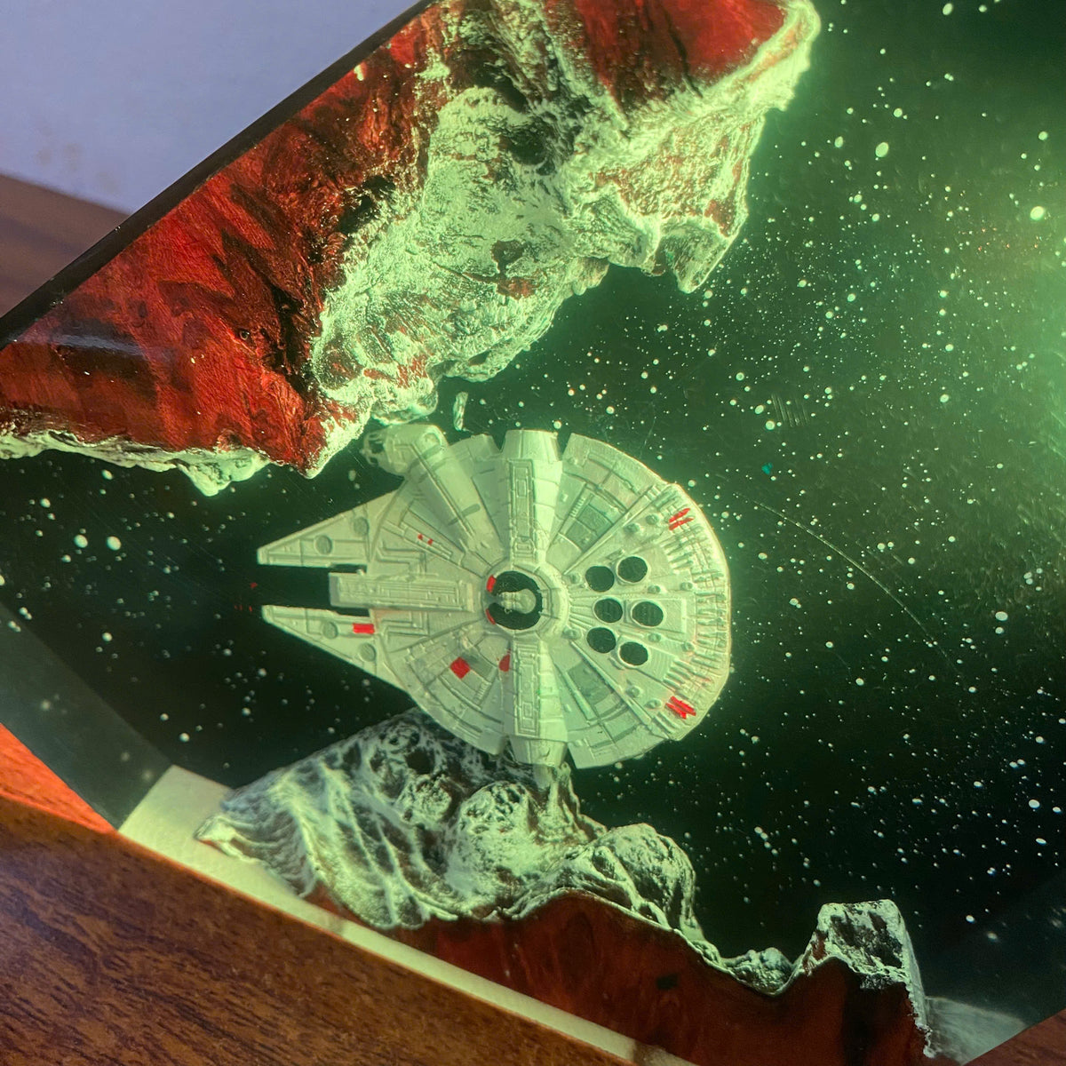 A 3D resin art piece depicting the Millennium Falcon flying between red and green rocky asteroids in space, with a wooden base visible at the bottom.