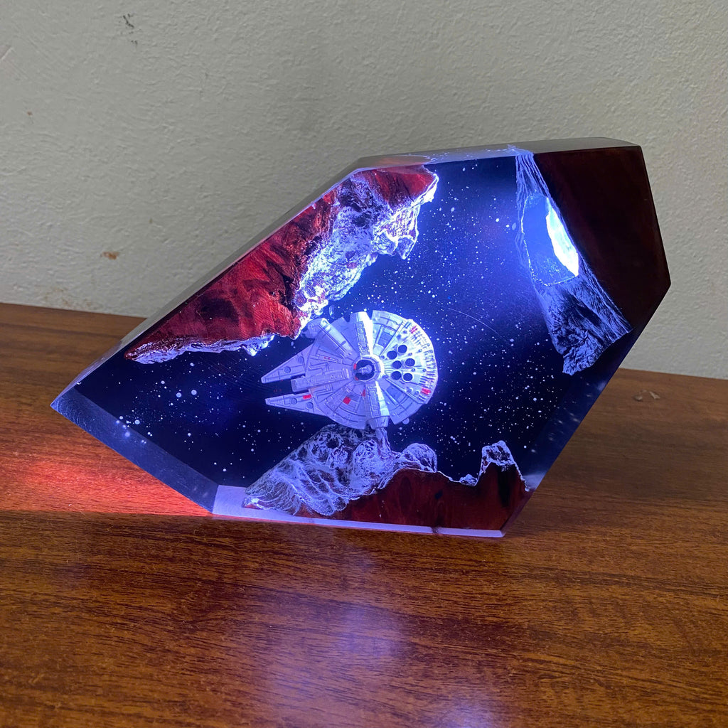 A faceted crystal or resin decorative object displaying a miniature space scene with the Millennium Falcon, glowing blue starry background, and red/orange rock formations, resting on a wooden surface against a light wall.
