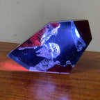 A faceted crystal or resin decorative object displaying a miniature space scene with the Millennium Falcon, glowing blue starry background, and red/orange rock formations, resting on a wooden surface against a light wall.