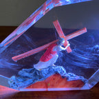 A clear resin geometric box containing a figurine of Jesus carrying a wooden cross, with swirling blue and red light reflections inside, resting on a dark wooden surface.