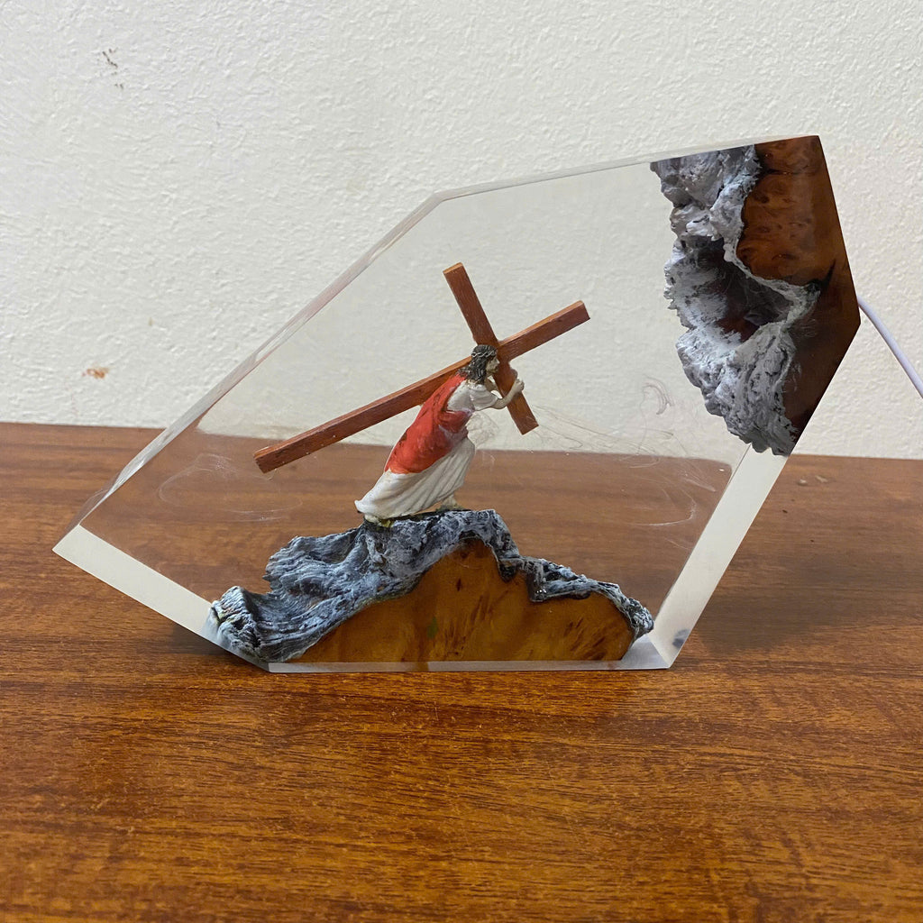 A clear acrylic display block containing a small figurine of Jesus carrying a wooden cross, set on a textured gray and gold base, displayed on a wooden surface against a white wall.