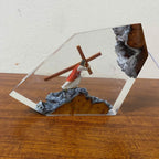 A clear acrylic display block containing a small figurine of Jesus carrying a wooden cross, set on a textured gray and gold base, displayed on a wooden surface against a white wall.