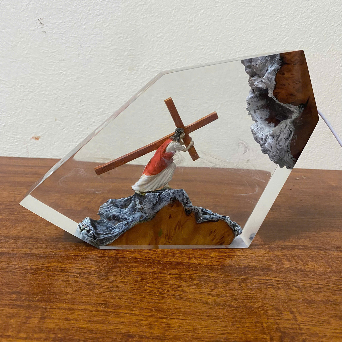 A clear acrylic display block containing a small figurine of Jesus carrying a wooden cross, set on a textured gray and gold base, displayed on a wooden surface against a white wall.