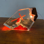A clear resin decorative plaque or lamp featuring a red cross, a robed figure, and textured stone-like details, placed on a wooden surface against a light blue wall. The piece is lit from within, casting red and warm light onto the tabletop.