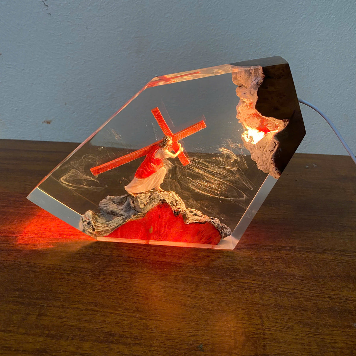 A clear resin decorative plaque or lamp featuring a red cross, a robed figure, and textured stone-like details, placed on a wooden surface against a light blue wall. The piece is lit from within, casting red and warm light onto the tabletop.
