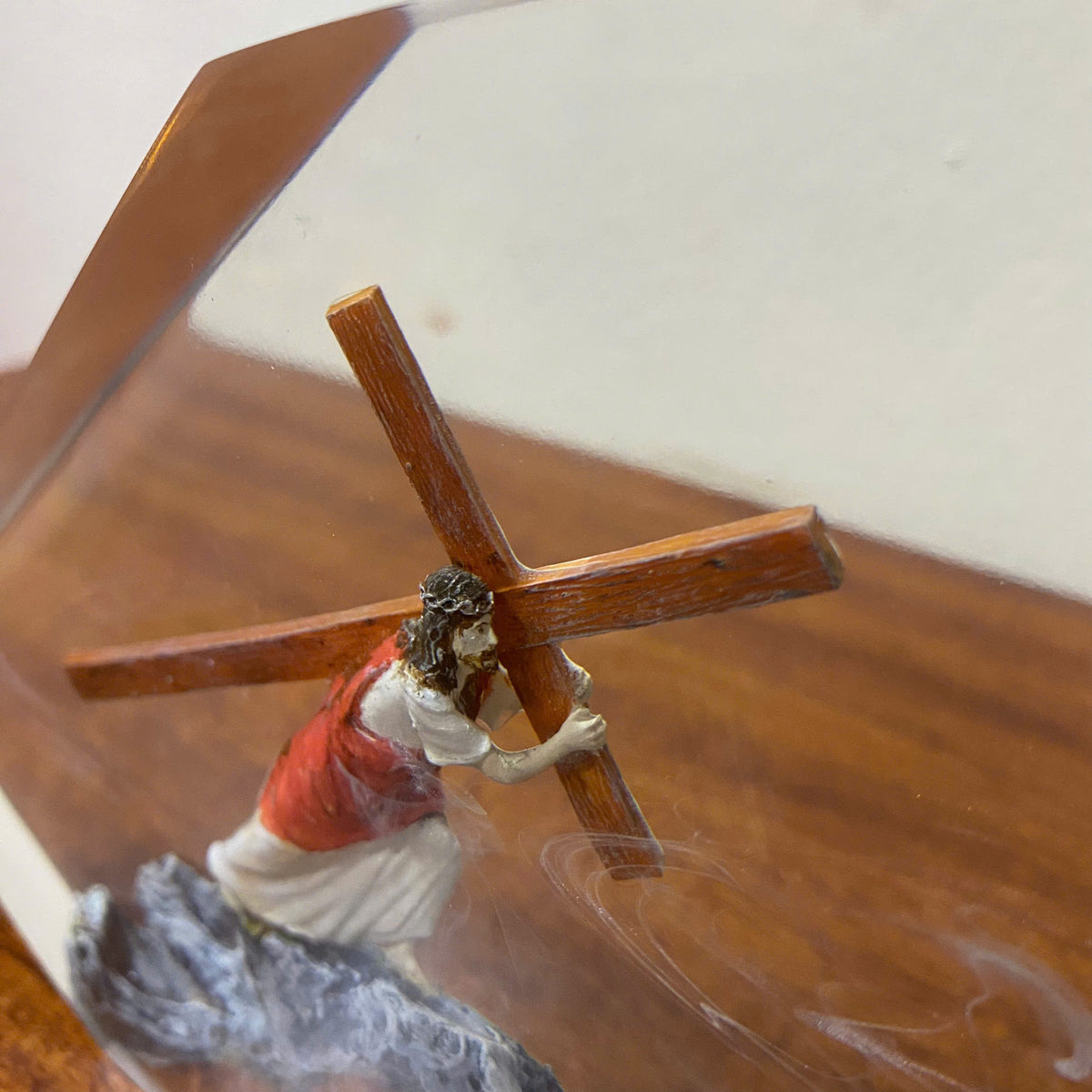 A small figurine of Jesus carrying a wooden cross, with a red and white robe and a rocky base, photographed close up against a neutral background.