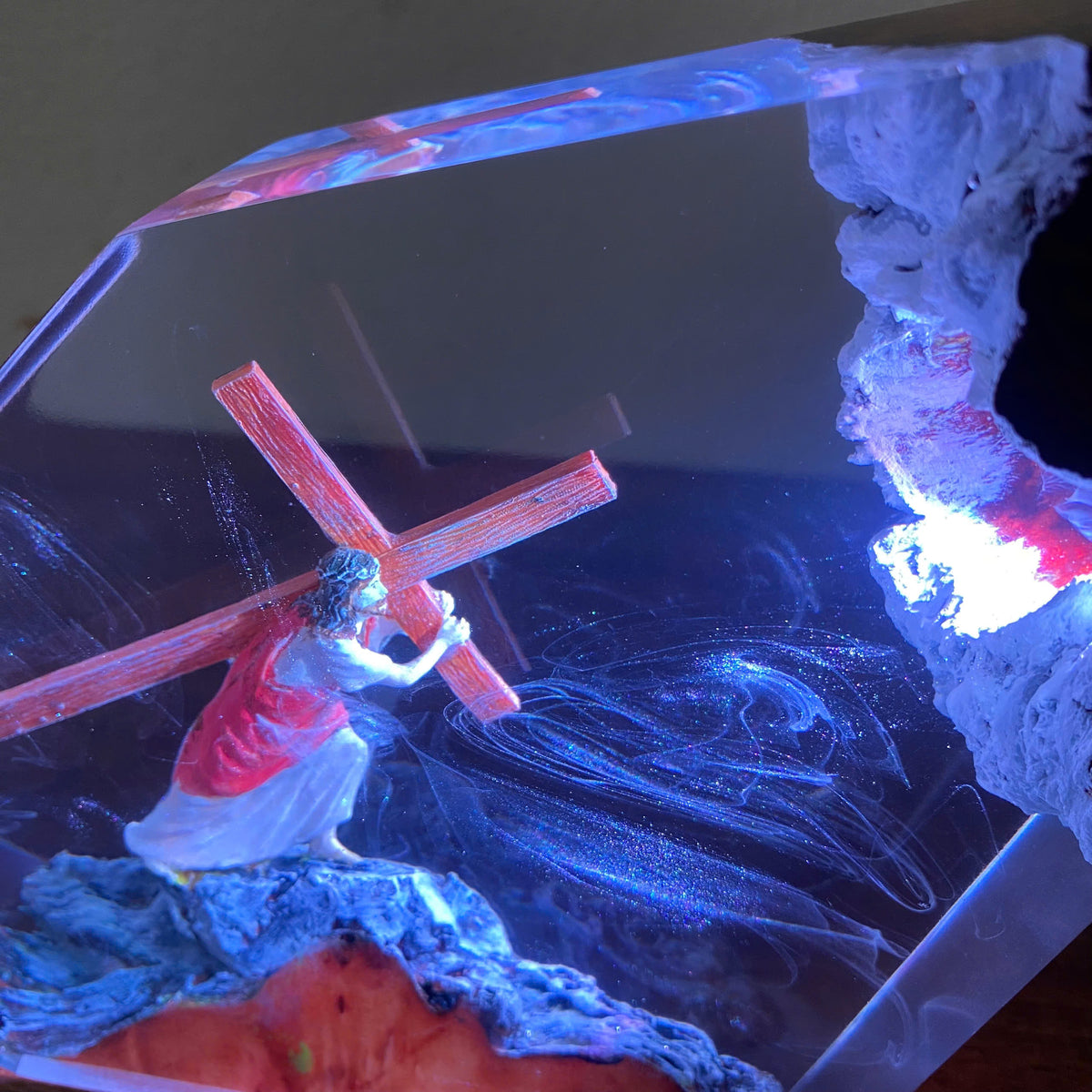 A clear resin decorative object featuring a small figurine of a man carrying a red cross, with swirling blue and white abstract patterns inside and a textured white rock-like form on the right side.