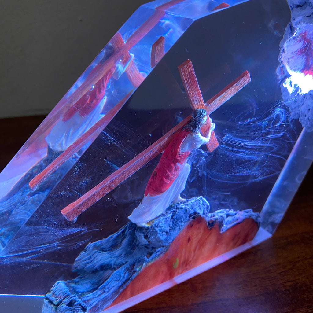 A resin crystal lamp featuring a miniature figure of Jesus carrying a wooden cross, set against a glowing blue and red background with a textured base.