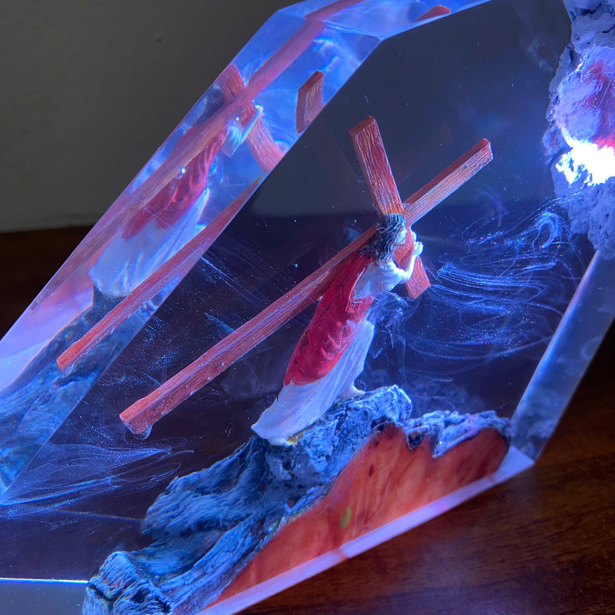 A resin crystal lamp featuring a miniature figure of Jesus carrying a wooden cross, set against a glowing blue and red background with a textured base.