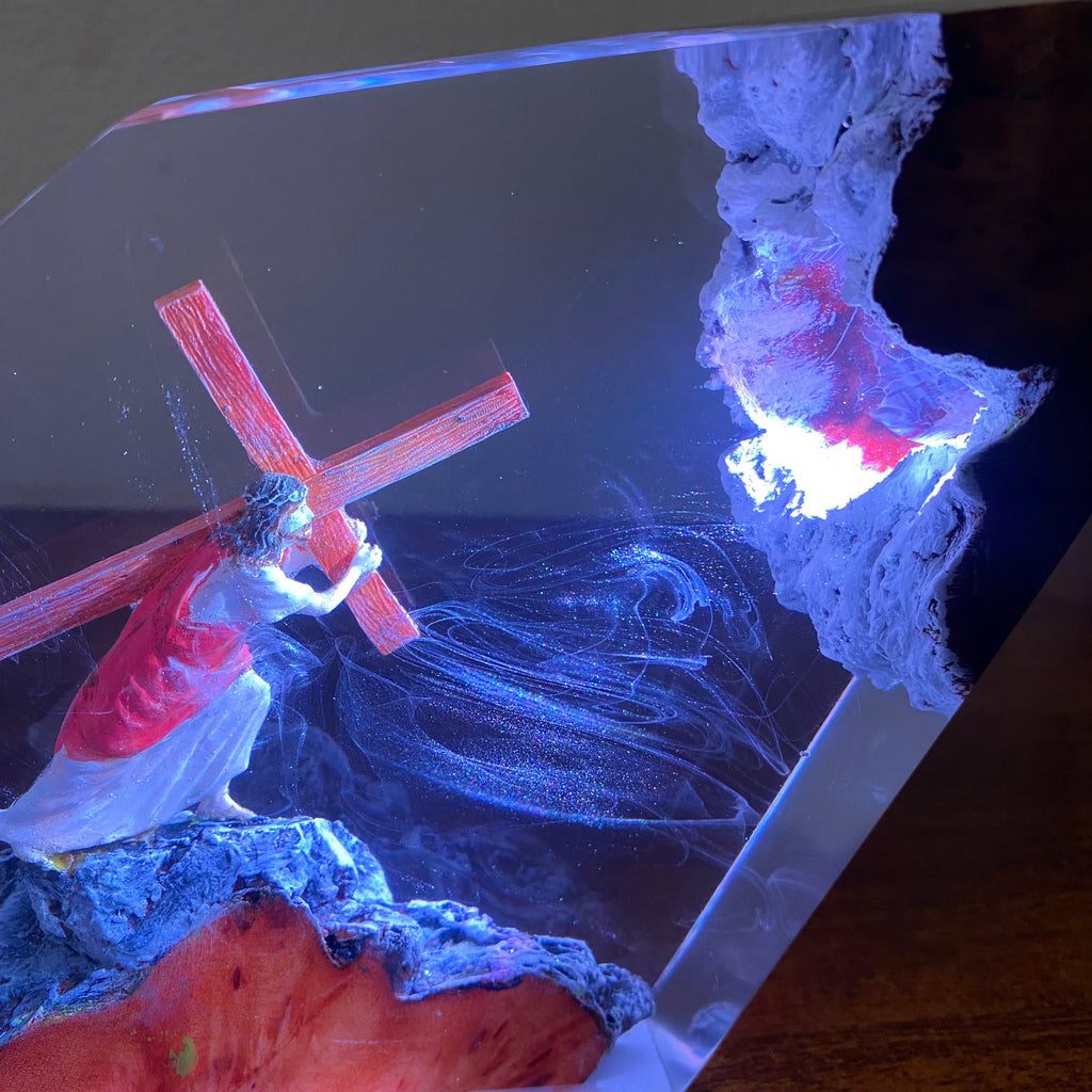 A clear acrylic or resin decorative piece featuring a sculpted figure carrying a red cross, with blue and purple glowing light effects inside the transparent panel and a textured white rock formation on the right.